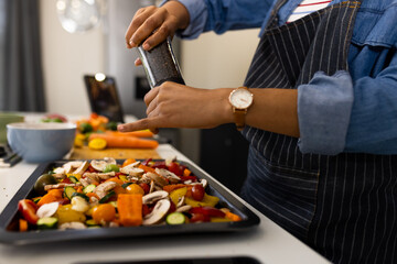 Midsection of biracial woman in apron preparing food in kitchen seasoning vegetables