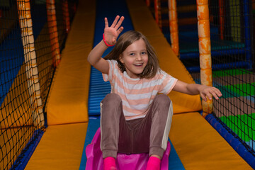 A little cheerful blonde girl slides down a slide in a children's entertainment center - 725397892