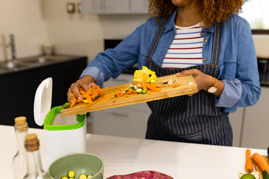 Midsection of biracial woman in apron composting vegetable waste in kitchen
