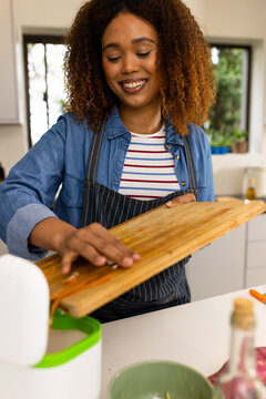 Smiling biracial woman in apron composting vegetable waste in kitchen