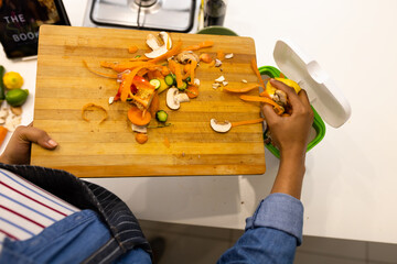 Overhead view of biracial woman in apron composting vegetable waste in kitchen