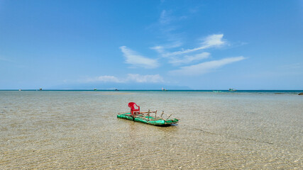 A serene beach scene with a makeshift raft and solitary red chair on clear shallow waters under a blue sky, suggestive of tranquility or vacation concept