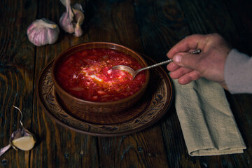 National Ukrainian borscht, earthenware, borscht with sour cream in a plate, a human hand scoops borscht with a spoon from a plate, garlic, napkin on a wooden table