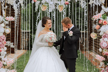 The groom cries during the wedding ceremony near the flower arch. Summer wedding in nature. She...