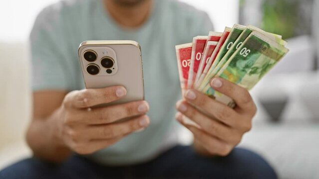 Young man relaxing on sofa at home, managing his wealth, counting shekels, texting business deals on smartphone