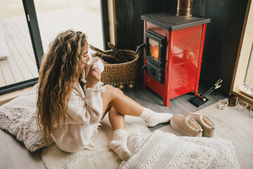 Young woman sitting by the fireplace in white sweater, drinking tea in cozy log cabin.