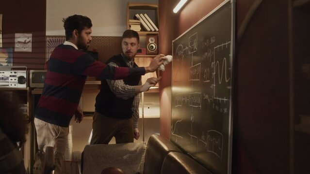 Medium shot of two multiethnic program developers working on engineering scheme together drawing it on chalkboard, thinking of creating startup in their old-fashioned garage style handmade office