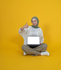 Young caucasian muslim girl sit floor legs crossed hold use laptop pc computer with blank screen mock up show thumb up like approve gesture. Isolated yellow studio background copy space. Advert idea.