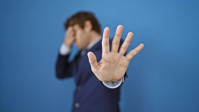 Terrified young man in business suit covers eyes, negating fear with stop gesture on isolated blue background. portrait of a desperate guy showing the sad reality of work stress.