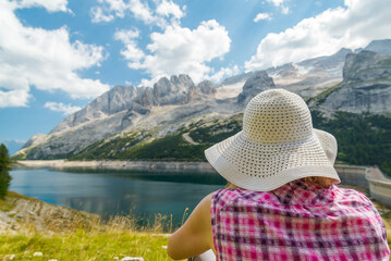 Young woman with fashionable white hat relaxing under the sun at the famous lake named "Fedaia", in the Dolomiti mountain chain, Italy. Marmolada mountain on the background.
