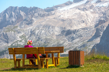 Blonde woman relaxing on a wooden bench on the grass. On the background the famous mountain named "Marmolada", in Dolomites mountain chain, Italy.