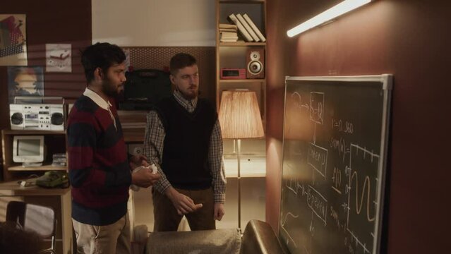 Medium shot of two ethnically diverse software engineers drawing program scheme on blackboard and discussing formulas while co-working in old-fashioned 90s style garage office