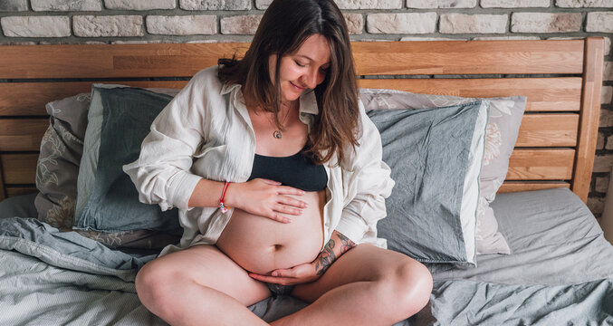 Young pregnant woman sitting with cross-legged tender touching belly on the bed in bedroom in the early morning time. Women's health, happy pregnancy and calm mental mood concept image.