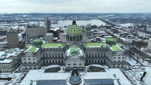 Pennsylvania capitol building covered in snow in downtown Harrisburg, PA after winter snowfall. Aerial reveal with Susquehanna River in background.