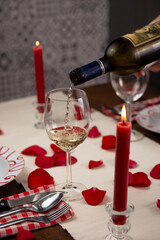 Hand serving a glass of white wine, on a dinner table set for two with red roses' petals. There is a lighted red candle. Vertical image