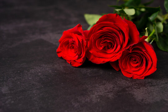 Set of three red roses in a bouquet laying on a dark grey textured surface. horizontal image