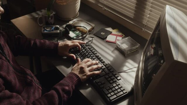 From above cropped shot of unrecognizable male programmer sitting at desk and typing on wired keyboard while working on old computer at home office