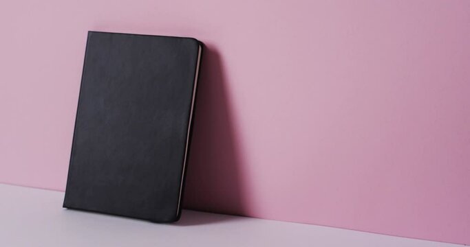 Close up of closed black book leaning on wall with copy space on pink background in slow motion