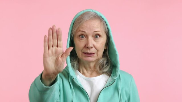 A serious-looking senior lady in a green hoodie with a raised hand showing the stop sign, posed against a pink backdrop. Emphasizing boundaries and caution. Camera 8K RAW.