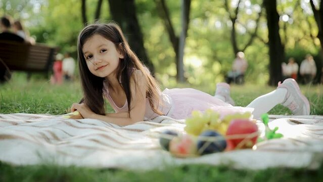 Weekend at picnic. Lovely caucasian girl on green grass meadow with basket full of fruits. Female child kid in pink dress on nature sit on white blanket having fun. Sunny summer