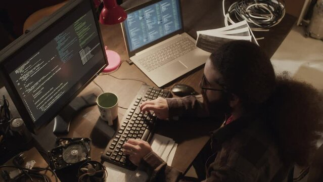 From above shot of long haired Middle Eastern hacker typing program code on old computer while working alone in dark old-fashioned room with dimmed warm yellow light