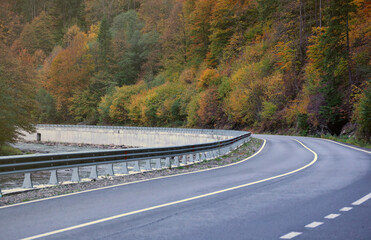 Road in autumn forest at sunset in Carpathian mountains, Ukraine. Beautiful mountain roadway with orange tress and high rocks. Landscape with empty highway through the woods in fall. Transportation