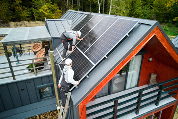 Technicians building photovoltaic solar module station on roof of house. Men roofers in helmets installing solar panel system outdoors. Concept of alternative and renewable energy. Aerial view. - obrazy, fototapety, plakaty