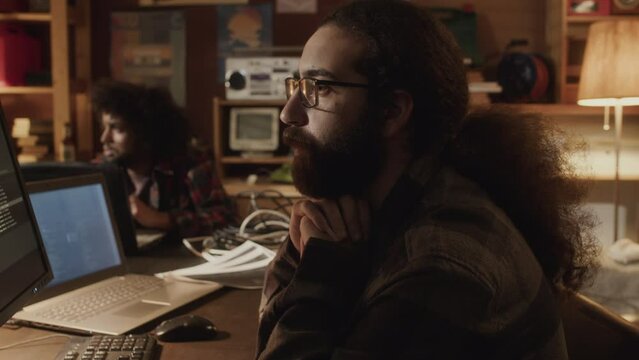 Waist up of Middle Eastern bearded tech lover with long hair, eyeglasses and vintage checkered shirt program coding on computer in dark handmade office with dimmed light