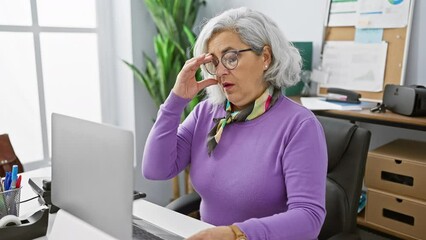 Mature woman working tiredly at computer in modern office, showing discomfort and stress.