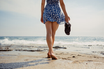 A woman enjoys a relaxed walk on the beach, holding her sandals, feeling the summer breeze and sea...