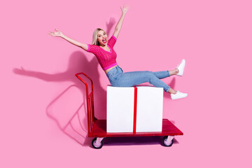 Full length photo of satisfied cheerful woman riding on shopping cart with large present raising...