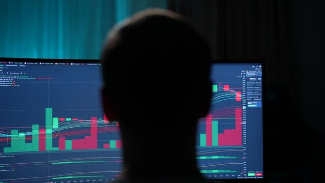Closeup view from shoulder of focused male stock trader sitting in front of computer monitor, making professional analysis of candlestick chart, creating strategy, looking at screen in dark room.