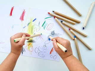 small child draws with colored pencils on paper on white table.