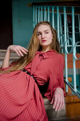Tall slender caucasian young woman with blond hair in coral dress sitting on concrete stairs of old...