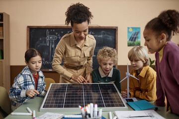 Diverse group of children learning about renewable energy in school with young teacher sowing solar panel to class - obrazy, fototapety, plakaty