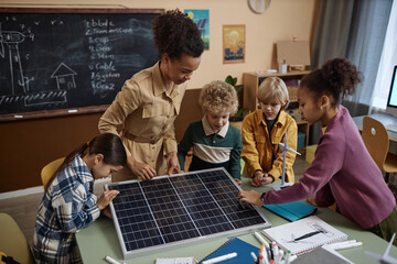 Multiethnic group of kids learning about renewable energy in school with young teacher sowing solar panel to class