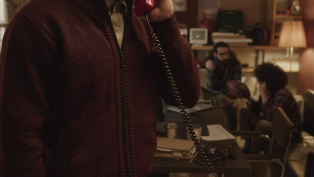 Tilt up shot of young Biracial guy dialing number on red vintage telephone while working on tech startup wit mates in old-fashioned apartment filled with technology gadgets and devices