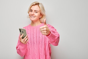 Fototapeta premium Portrait of young blond woman in pink sweater holding mobile phone in hand with happy smiling face. Person with smartphone isolated on white background.