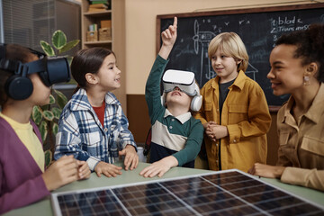Diverse group of children in modern school classroom with little boy wearing VR headset and pointing up enjoying immersive technology