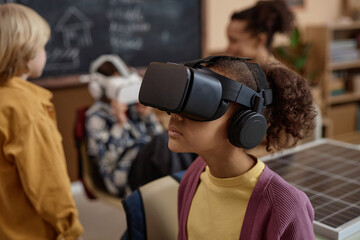 Side view portrait of young Black girl wearing VR headset in class and enjoying immersive learning...