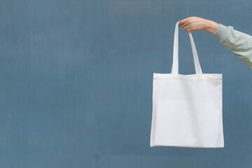 Blank white tote bag canvas fabric with handle mock up design. Close up of woman hand holding eco or reusable shopping bag on blue metal wall. No plastic bag and ecology concept.