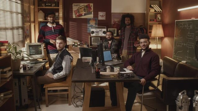 Slowmo portrait of ethnically diverse team of male geeks posing for camera in old-fashioned 90s style apartment filled with tech details and gadgets