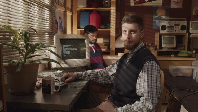 Medium portrait of Caucasian young geek wearing vintage checkered shirt and knitted vest posing for camera sitting at desk in front of old computer monitor with program code on display