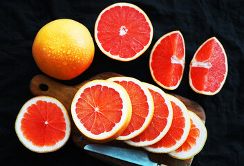 Sliced ​​and whole grapefruit on a wooden board next to a knife on a dark background