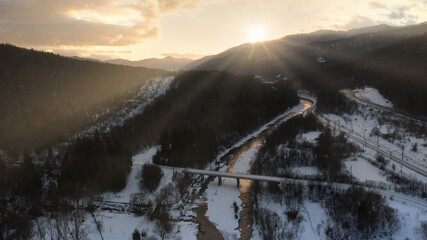 Winter sunrise over bridge crossing partially frozen river with surrounding snow-covered landscape.