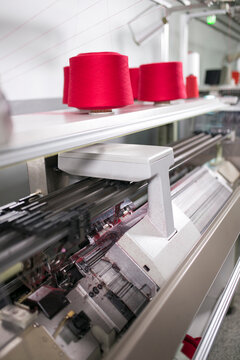 Automated flat knitting machine manufacturing textile with red yarn spools in a factory.
