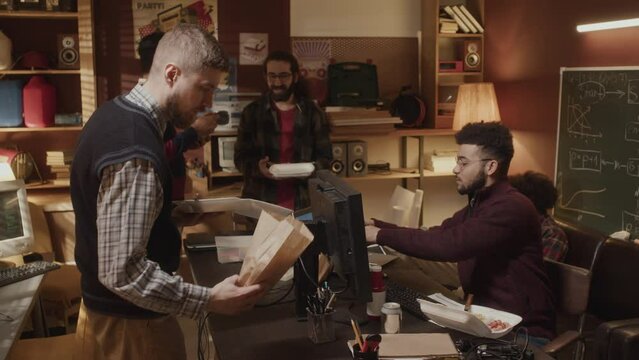 Medium shot of five young male ethnically diverse software startupers having lunch together while co-working on their tech business in 90-s style apartment - Powered by Adobe