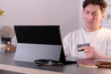 Dissapointed young man with a laptop and credit card in hand, passport and wallet on the table
