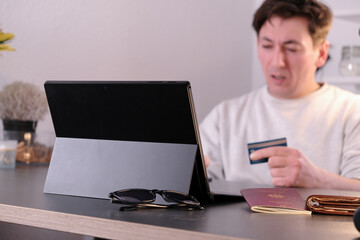 Shocked young man with a laptop and credit card in hand, passport and wallet on the table