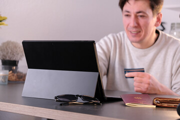 Pleased happy young man with a laptop and credit card in hand, passport and wallet on the table